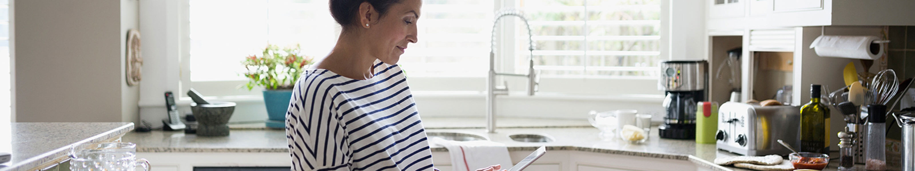 A woman is standing in her kitchen and looking at a mobile tablet.