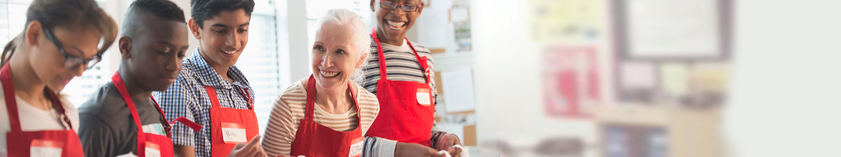 Young teens and older woman are wearing red aprons in a kitchen.