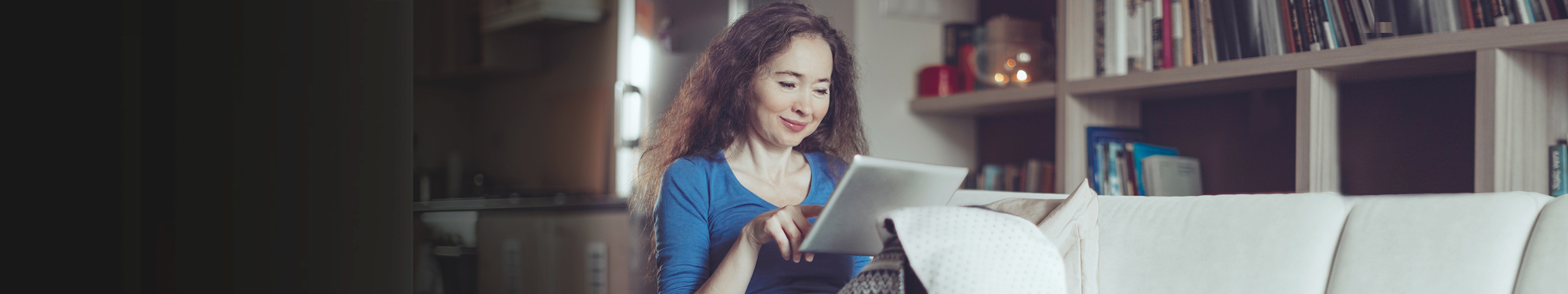A middle-aged woman sits comfortably on her sofa while surfing on her mobile tablet.